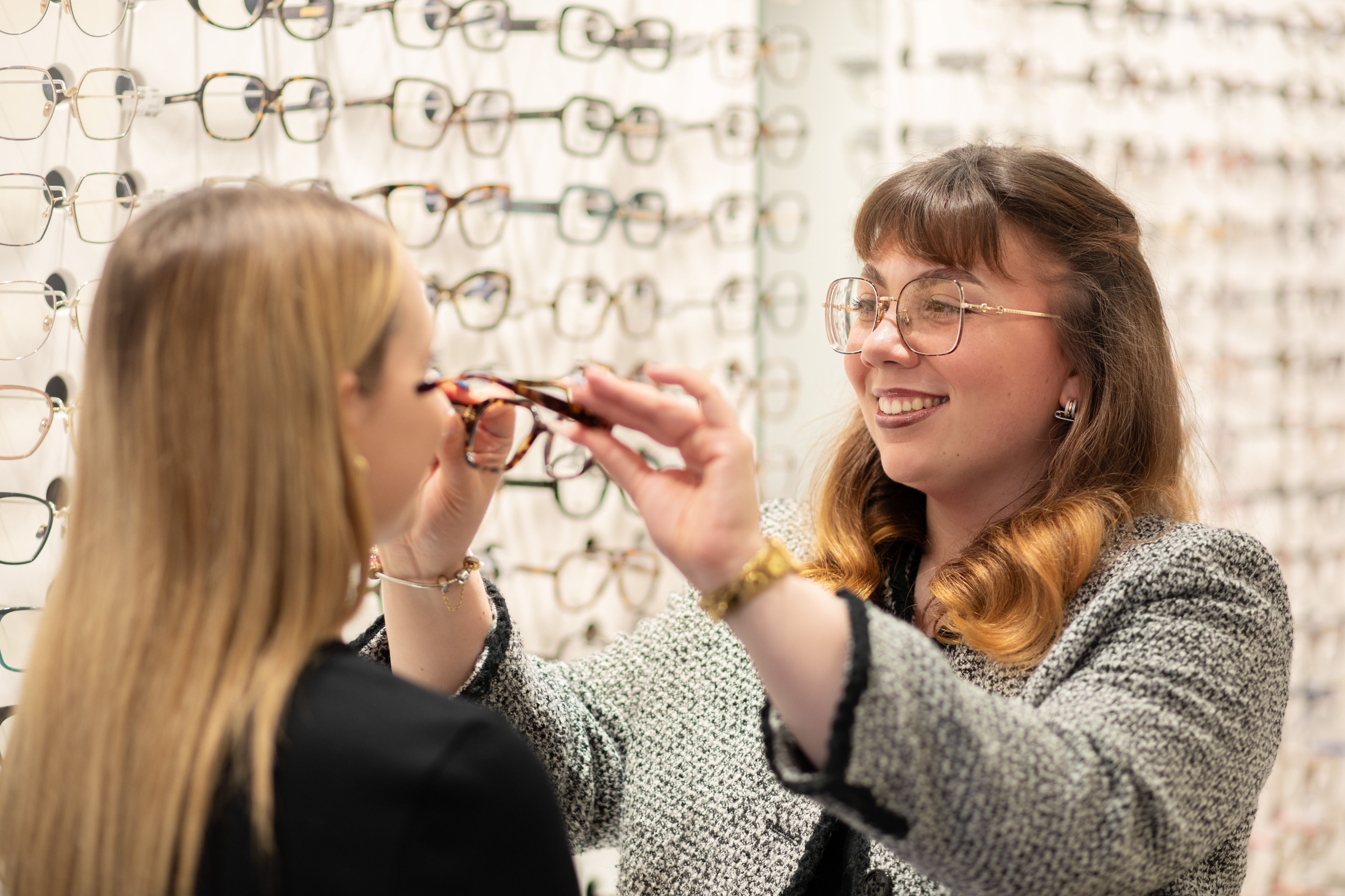 une opticienne s'apprête à mettre une monture de lunettes sur le nez d'une cliente devant un facing de lunettes